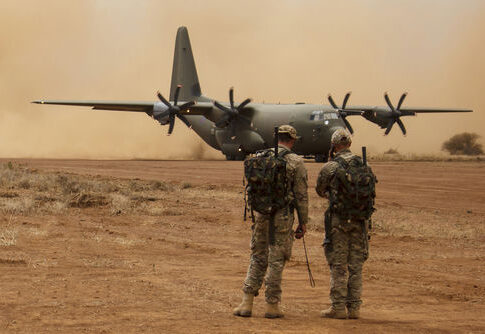 Two soldiers watch a military transport plane on a dusty airstrip
