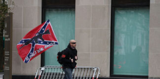 Man walking with Confederate flag on city sidewalk.