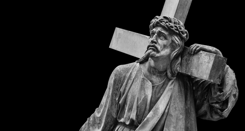 shutterstock_1937672293.jpg A black and white statue of Christ carrying a cross with a sorrowful expression
