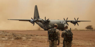 Two soldiers watch a military transport plane on a dusty airstrip